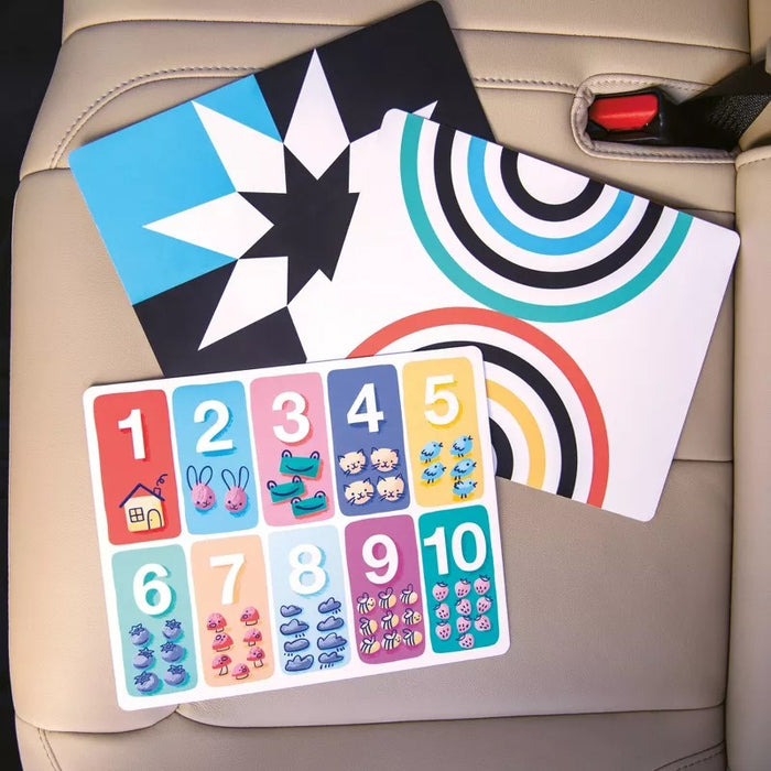 Three colorful educational cards rest on the beige Nuby Safe in the Seat Car Seat Protector: one with stars, one with curved stripes, and one showing numbers 1–10 with animals and objects. Perfect for both rear- and forward-facing car seats.