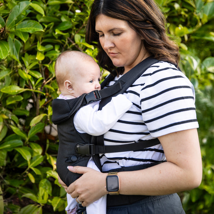 A woman gazes down at her baby, who looks to the side, as she stands outdoors in front of green bushes wearing a striped shirt and using the Nuby SureFit Baby Carrier.