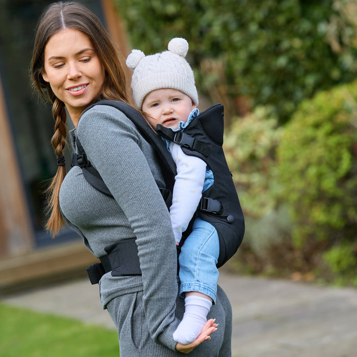 A woman in gray carries a baby on her back in the SureFit Baby Carrier. The baby, wearing light blue jeans, a gray sweater, and a white pom-pom hat, looks at the camera as they spend time outdoors in a garden.
