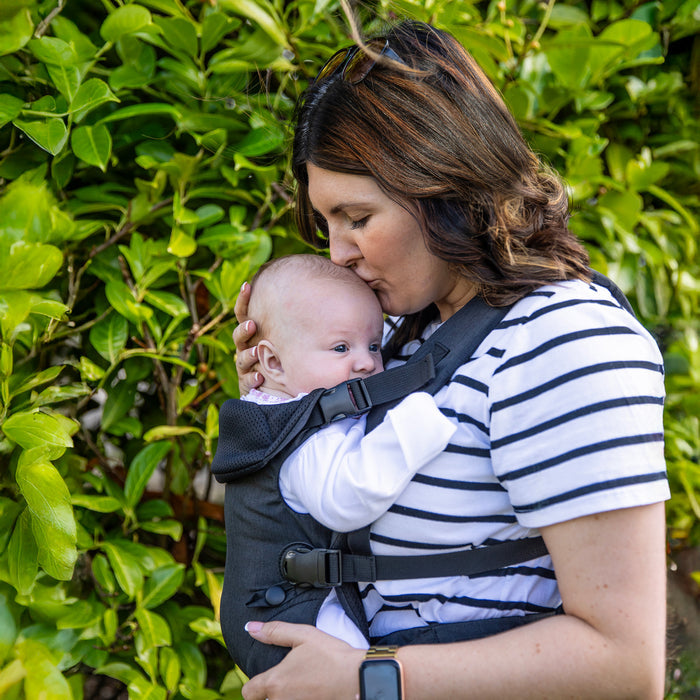 A woman in a striped shirt kisses a baby on the forehead while carrying the baby in the Nuby SureFit Baby Carrier, standing in front of green leafy bushes.