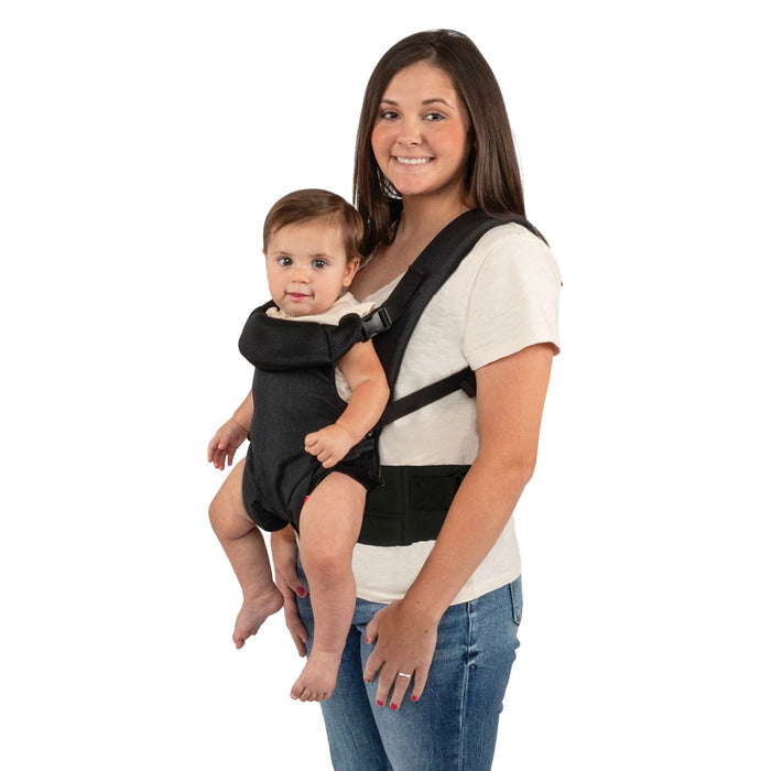 A woman smiles while carrying a baby in the Nuby SureFit Baby Carrier, wearing a white t-shirt and jeans against a white background.