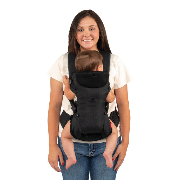 A woman smiles while standing and wearing the Nuby SureFit Baby Carrier with her baby facing inward on her chest. The baby’s arms and legs are visible, and both look relaxed and comfortable in this adjustable carrier.